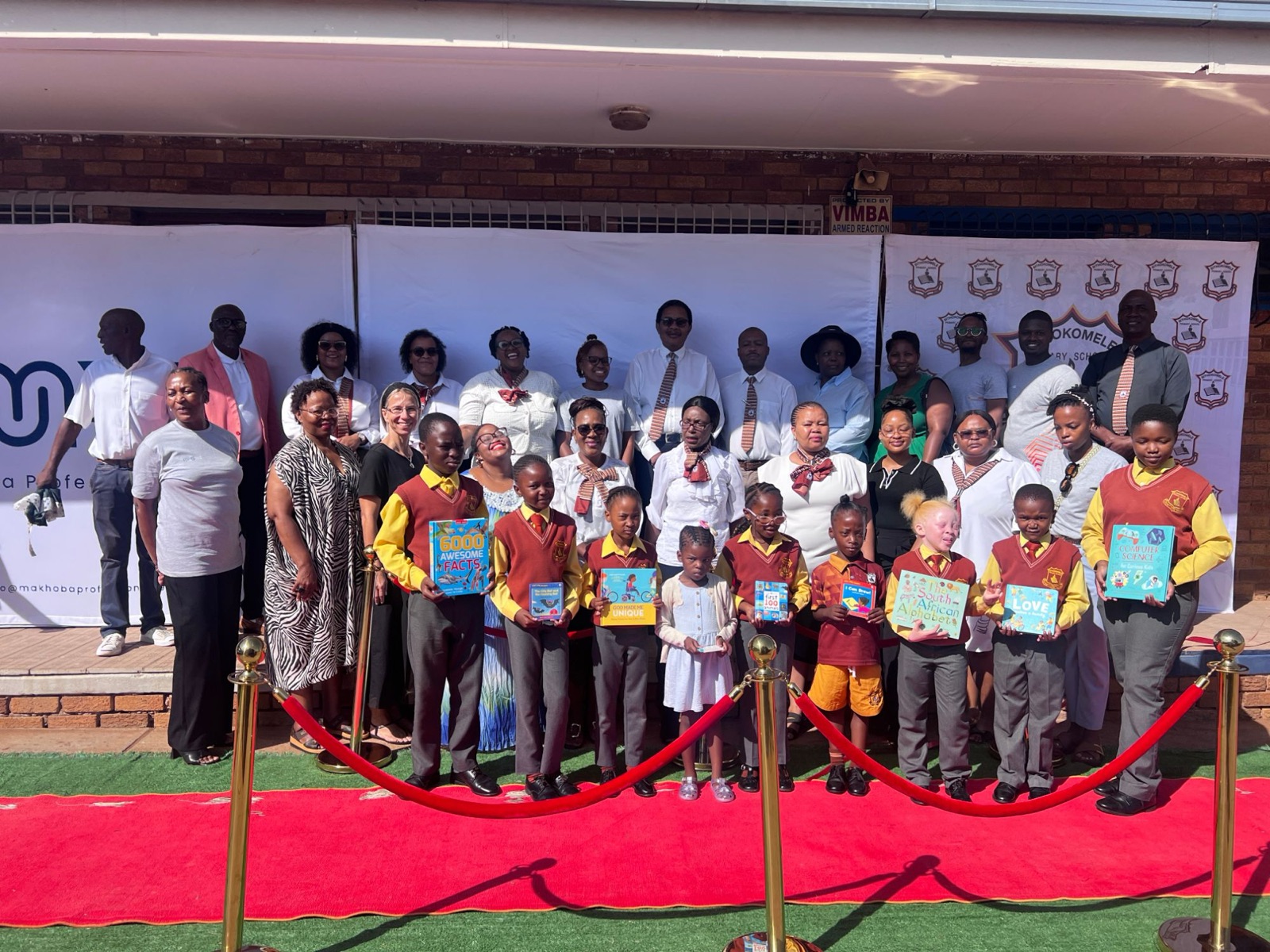 Learners holding donated books stand on a red carpet with teachers, partners and the Shabbos Shalom team
